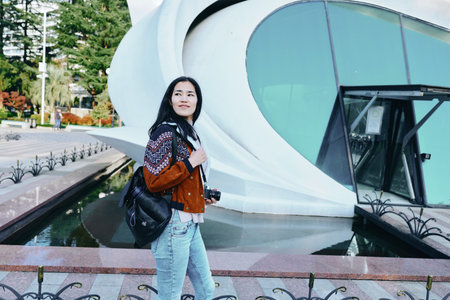 Woman with backpack posing beside futuristic building, casual fashion, outdoor travel scene, modern architecture, daylight urban environment, exploration and discovery.の写真素材