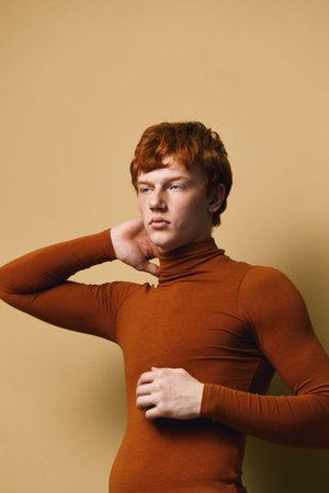 Young man with red hair posing in brown turtleneck sweater against beige background with thoughtful gaze and hand touching neck in studio portrait.の写真素材