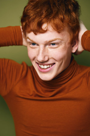 Young man with red hair smiling joyfully wearing brown turtleneck sweater standing against green background. Casual portrait of happy person with hands behind head.の写真素材