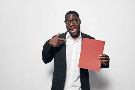 African American man with dark skin wearing glasses and black jacket pointing at red paper sheet on white background. Confident male holding blank document for presentation or advertisement.の写真素材