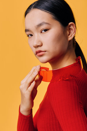 Young woman portrait with long dark hair wearing red turtleneck sweater and holding red glasses near face on orange background for fashion and style concept.の写真素材