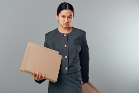 Young woman in dark business suit holding cardboard boxes looking serious in studio, isolated on gray background for delivery, logistics, or packaging concept.の写真素材