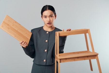 Woman holding broken wooden chair and detached plank indoors, surprise and concern on face, furniture damage repair concept with casual outfit on gray background.の写真素材