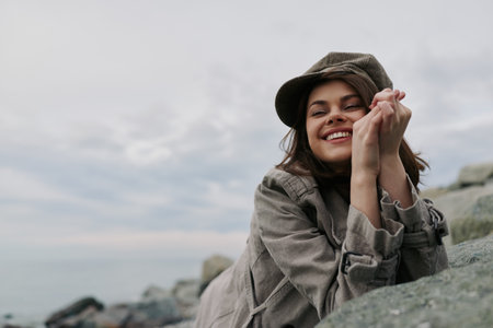 Smiling young woman in casual clothing and hat rests on rocks near calm sea under cloudy sky. Natural outdoor scene with relaxed mood and soft natural light, lifestyle and happiness concept.の写真素材