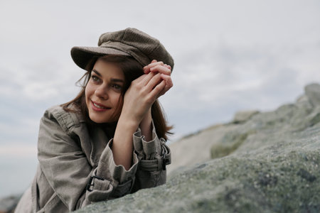 Smiling young woman in vintage hat and coat leans on rock outdoors with cloudy sky in background. Concept of autumn fashion, nature, and relaxed lifestyle in soft natural light.の写真素材