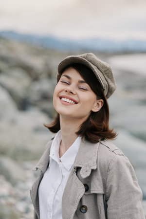 Smiling young woman in casual beige jacket and hat enjoys outdoor nature moment. Soft light highlights relaxed mood and natural beauty with blurred rocky background.の写真素材