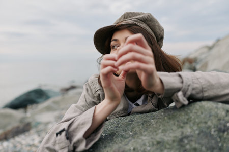 A thoughtful young woman wearing a tweed cap leans on a large rock outdoors, gazing into the...