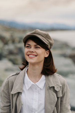Young woman wearing a hat and coat smiles outdoors near water and rocks. Natural light highlights her joyful expression and casual autumn outfit. Concept of happiness and nature.の写真素材