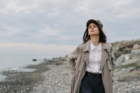 Young woman in trendy coat and hat stands on rocky beach with cloudy sky. She looks confident and thoughtful, with hands in pockets and natural makeup. Outdoor autumn fashion concept.の写真素材