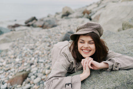 Smiling young woman in casual clothes and hat lies on rocky beach, looking away with relaxed expression. Concept of leisure, nature, and happiness outdoors by the sea.の写真素材