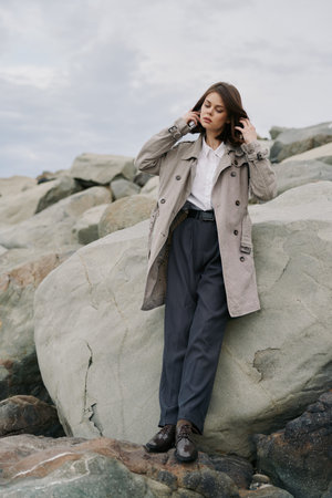 Young woman in stylish coat and trousers stands on large rocks outdoors under cloudy sky. Fashion...