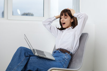 A relaxed young woman sits in a modern chair by a bright window, yawning and stretching after a long...