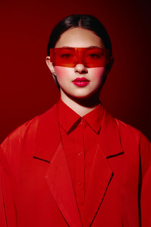 Young woman wears oversized red sunglasses and bold red lipstick in a red blazer against a red background. Creative fashion portrait focusing on beauty and style.の写真素材