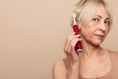 Mature woman with blonde hair holds red electric toothbrush near face on beige background. Concept of oral hygiene, dental care, personal grooming, and healthy lifestyle.の写真素材