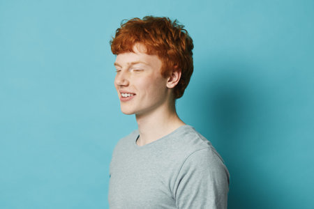 Young man with red hair and freckles wearing gray t shirt standing with eyes closed against blue background. Casual portrait of a smiling male model in studio with soft lighting.の写真素材