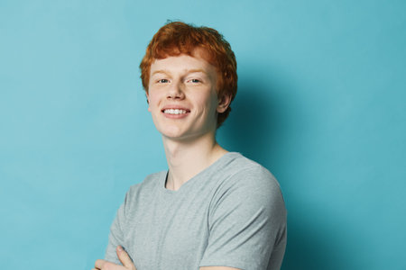 Young man with red hair smiling and crossed arms in casual t shirt against plain blue background. Portrait of confident male with light skin and curly hair studio shot.の写真素材