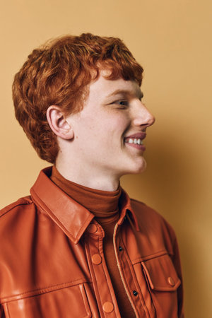 Young man with red hair wearing brown leather jacket and turtleneck smiling in profile against beige background. Curly hairstyle and casual fashion with happy expression and natural light.の写真素材