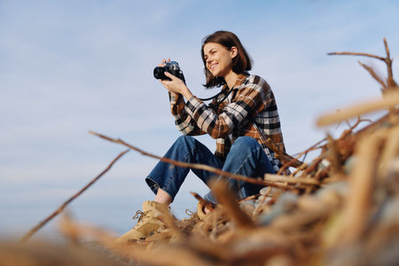 Photographer woman outdoors on a sandy beach, seated on driftwood, holding a camera and capturing...