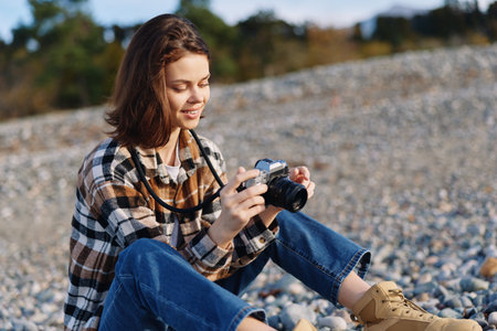 Photographer woman outdoors, sitting on pebbles with a camera, wearing a plaid shirt and jeans, capturing coastal landscape and natural light. An intimate, relaxed moment in open scenery, perfect forの写真素材