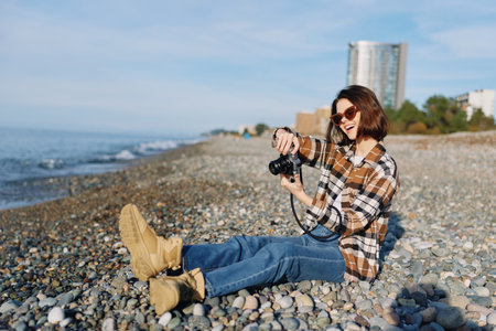 Photographer sitting on a pebble beach with a camera, wearing a plaid shirt, jeans and boots, enjoying a sunny day by the sea with distant buildings and a blue sky.の写真素材