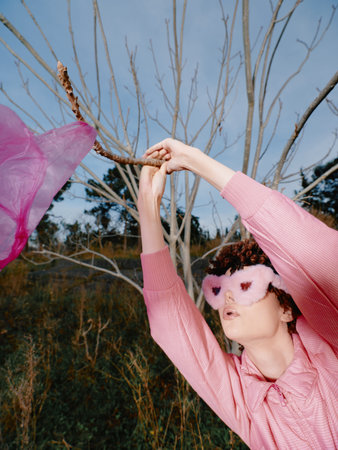 Woman in a pink sweater wearing a pink eye mask dances outdoors among bare trees, capturing a playful whimsical moment with flowing fabric and motion.の写真素材