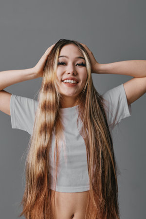 Young woman with long straight hair smiling and posing with hands on head studio portrait against gray background. Casual appearance wearing white cropped t shirt and relaxed expressionの写真素材