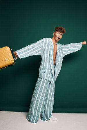 Young man in striped pajamas holding yellow suitcase with excited expression standing against green background. Casual travel, leisure, and vacation concept with comfortable clothing and luggage.の写真素材