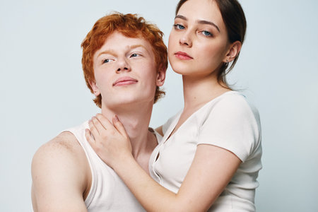 Couple posing portrait with woman embracing man tenderly, young adults in casual white tops looking confident, studio background with soft lighting.の写真素材