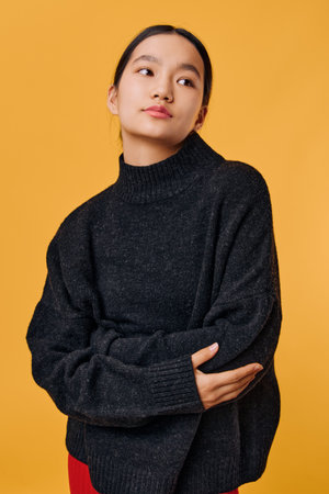 Young woman in black sweater poses with crossed arms against yellow background, looking to side with...