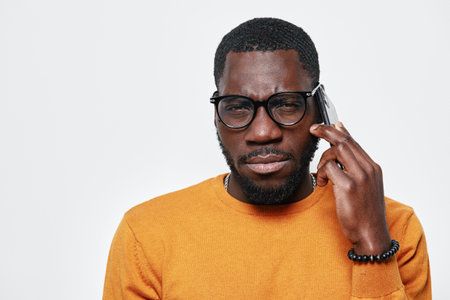 African American man with dark skin wearing glasses and orange sweater talking on smartphone with...