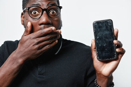 African American man with glasses showing shocked expression covering mouth holding smartphone with...