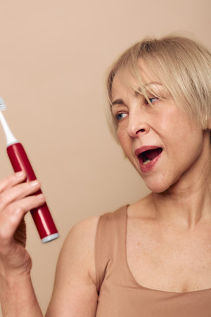 Middle aged woman with short blonde hair holds red electric toothbrush looking at it with open mouth. Concept for oral hygiene, dental care, personal routine, and healthy lifestyle.の写真素材