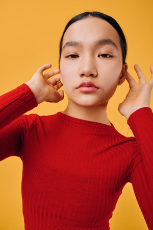 Young woman in red sweater posing with hands near ears on yellow background studio portrait. Asian...