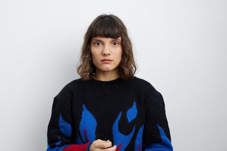 A thoughtful young woman stands against a plain wall wearing a dark sweater with blue flame patterns, conveying calm confidence and introspection in a studio portrait.の写真素材