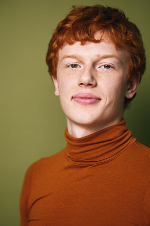 Portrait of young man with red hair and fair skin wearing burnt orange turtleneck looking confident with slight smile and neutral green background in studio lighting.の写真素材