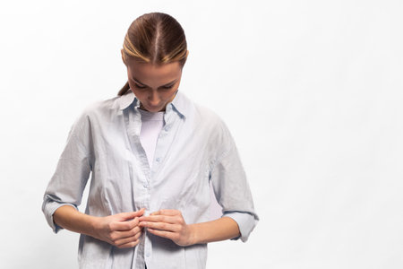 Young woman in casual shirt buttoning up, looking down. Minimalist studio on white background. Concept for clothing, fashion, daily routine, and casual style.の写真素材
