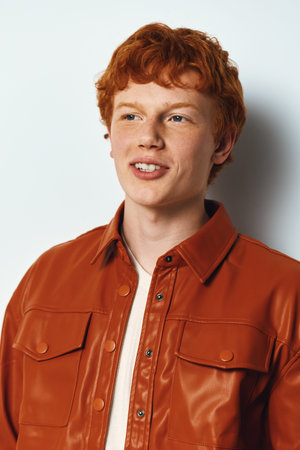 Young man with red hair and freckles wearing a brown leather jacket smiles looking away with white...