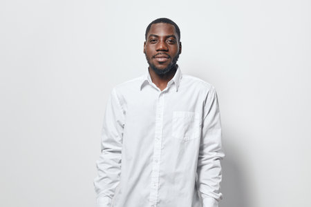 African American man with dark skin wearing white shirt standing against white background looking at camera with calm expression and relaxed posture in studio portrait.の写真素材