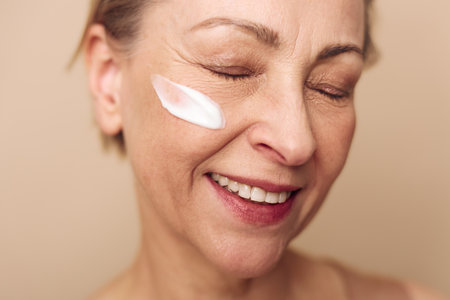 Close-up of mature woman with natural skin smiling with eyes closed while applying moisturizing cream on cheek. Concept of skincare, anti-aging, beauty, and facial care routine.の写真素材