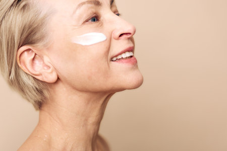 Smiling mature woman applies white face cream on cheek, showing skincare routine and healthy glowing skin. Concept of beauty, antiaging, wellness, selfcare, and natural skincare products.の写真素材