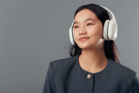 Young woman wearing wireless headphones and looking away with slight smile against plain gray background. Portrait of confident female in dark clothing with natural makeup and long hair for lifestyleの写真素材