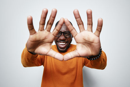 African American man with dark skin wearing glasses and orange sweater smiling and framing face with...