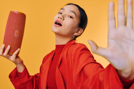 Woman in red jacket holding portable speaker while extending hand against yellow background. Girl...