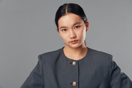 Portrait of confident Asian woman with natural makeup and dark hair tied back wearing a gray...