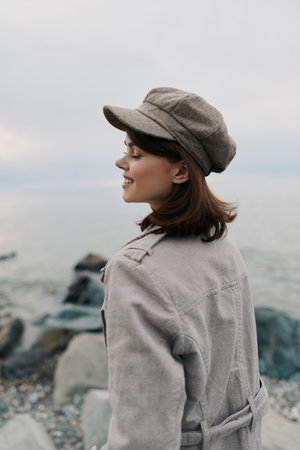 Young woman in a cozy coat and hat stands near rocky shore looking sideways, smiling gently....