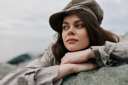 Thoughtful young woman wearing a cap leans on a rock outdoors. She looks to the side with a calm...