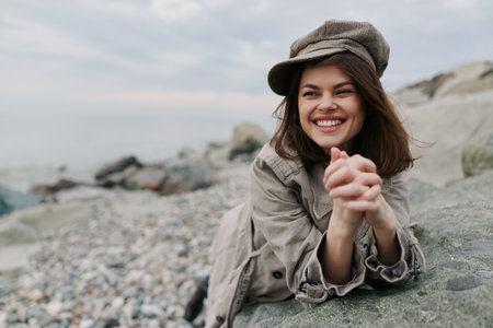 Smiling young woman in casual outfit and hat relaxing on rocky beach. She looks happy and carefree with natural background of stones and cloudy sky. Concept of outdoor leisure and joy.の写真素材