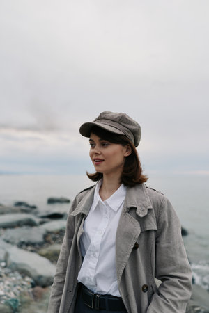 Young woman wearing gray coat and cap stands on rocky beach near calm sea. Overcast sky creates soft light and peaceful atmosphere. Concept of autumn fashion and outdoor portrait.の写真素材