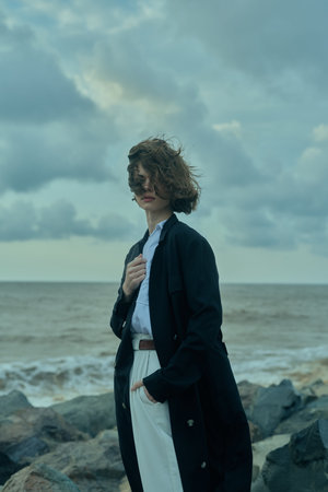 Windy seaside scene with a long dark coat on a rocky shore, windblown hair, moody sky, calm ocean,...