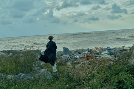 Woman standing on a rocky shore by the sea, gazing at waves, bag over shoulder, moody clouds, coastal landscape, solitude and reflectionの写真素材